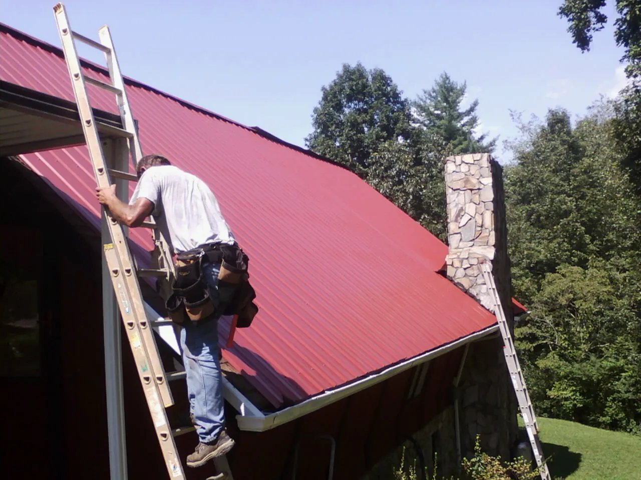Roofer on a ladder beside a red metal roof and stone chimney — Soffit & Fascia Repair by Roofing Brothers in East Millsboro